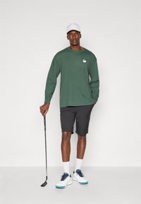 Hosszú ujjú zöld póló logóval, fekete sort, fehér sapka és kék részletekkel ellátott sportcipő, golflabdát tartva egy sima háttér előtt.