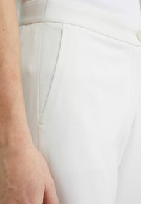 Pantaloni in misto cotone bianchi caratterizzati da una texture liscia, tasche laterali e una vestibilità su misura. Cuciture dettagliate sono visibili vicino alle tasche.