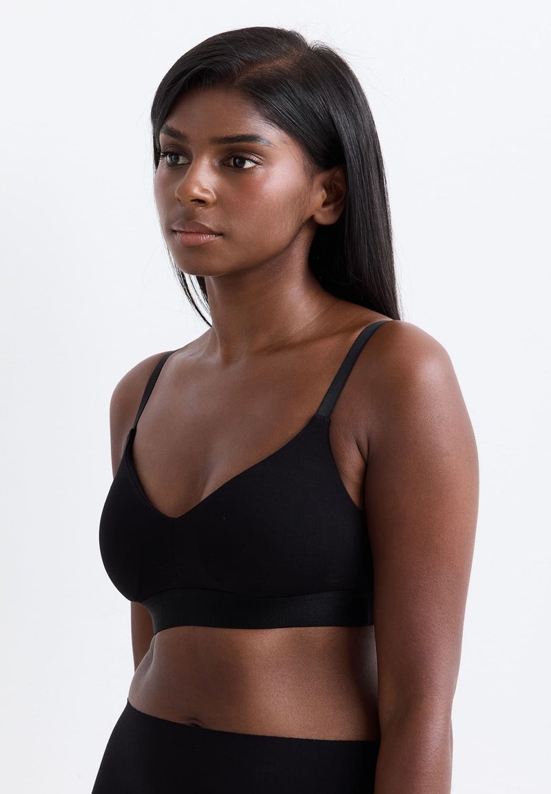 Junge Frau mit glattem, dunklem Haar, trägt ein schwarzes Bralette und eine schwarze Unterhose, steht links vor einem schlichten weißen Hintergrund.