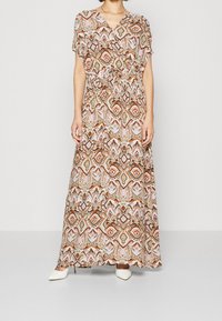 La robe maxi présente un motif géométrique dans des tons chauds, avec une taille froncée, des manches courtes et un tissu léger. Associée à des chaussures blanches.