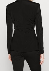 Černé přiléhavé sako s dlouhými rukávy, designem s přepínáním vzadu a hladkou texturou. Střižený střih s jemnými detailem na sbírání.