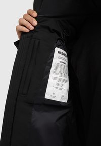 Interior de una chaqueta acolchada negra, con una etiqueta de tela que incluye instrucciones de cuidado, un bolsillo con cremallera y una textura suave y mate.