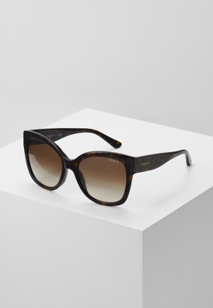 Tortoiseshell square sunglasses with gradient brown lenses and “vogue” logo on temples, resting on a white pedestal against a gray background.