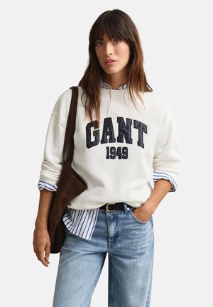Frau trägt weißen GANT-Sweatshirt mit der Aufschrift "1949", gestreiftes Hemd darunter, blaue Jeans, schwarzen Gürtel und braune Umhängetasche, steht allein.