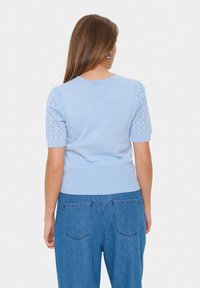 Light blue knitted sweater with short, puffed sleeves featuring a lacy pattern. Ribbed hem and smooth texture, paired with high-waisted denim.