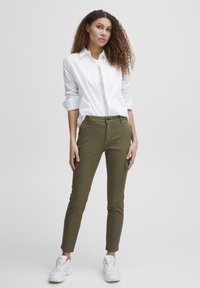 Pantalons cargo verts avec poches zippées, coupe fuselée, et en mélange de coton, associés à une chemise blanche à boutons et des baskets blanches.