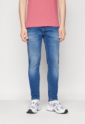 Personne portant un jean slim bleu, des chaussures de sport blanches et grises, et un t-shirt rose à manches courtes, debout devant un fond uni.
