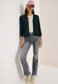 Donkergroene cardigan met knopen, beige t-shirt, grijze rechte jeans en lichte sneakers; staand tegen een neutrale achtergrond.