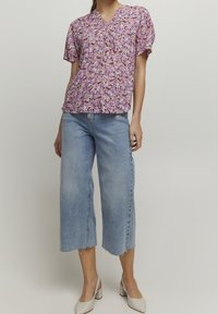 Blomstret kortærmet bluse i nuancer af lilla, med V-udskæring og samlede detaljer foran, parret med lyseblå cropped jeans.