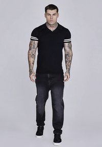 Černé polotriko s bílými pruhy na rukávech, v kombinaci s tmavými džínami a černými teniskami. Modelka ukazuje pohled zepředu, stojí.