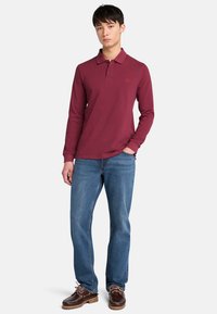 Mladý muž stojí s neutrálním výrazem, oblečený v vínově červeném polo tričku s dlouhým rukávem, modrých džínách a hnědých botách.