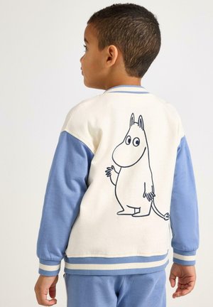 Bambino con giacca bianca e blu con disegno del personaggio Moomin sul retro, in piedi e che guarda di lato su uno sfondo neutro.
