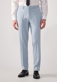 Pantalones azul claro de tejido suave, con pliegues, un ajuste entallado y bolsillos laterales. Combinados con zapatos de vestir negros.