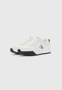 Witte Calvin Klein lage sneakers met zwarte zolen, veters en zijlogo, iets naar links gekanteld op een effen achtergrond.