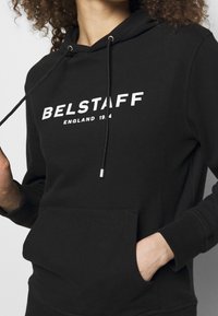 Svart hoodie med dragsko, framficka och vit "BELSTAFF" logotyp med texten "ENGLAND 1944". Mjuk tyg, avslappnad design.