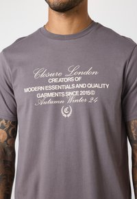 Graues Baumwoll-T-Shirt mit weißem Text, der "Closure London" und "Herbst Winter 24" zeigt, mit einem lässigen Schnitt und kurzen Ärmeln.
