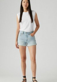 Top blanco sin mangas, pantalones cortos de mezclilla azul claro con puños enrollados, y sandalias negras con tiras. El modelo está de pie con la mano en el bolsillo.