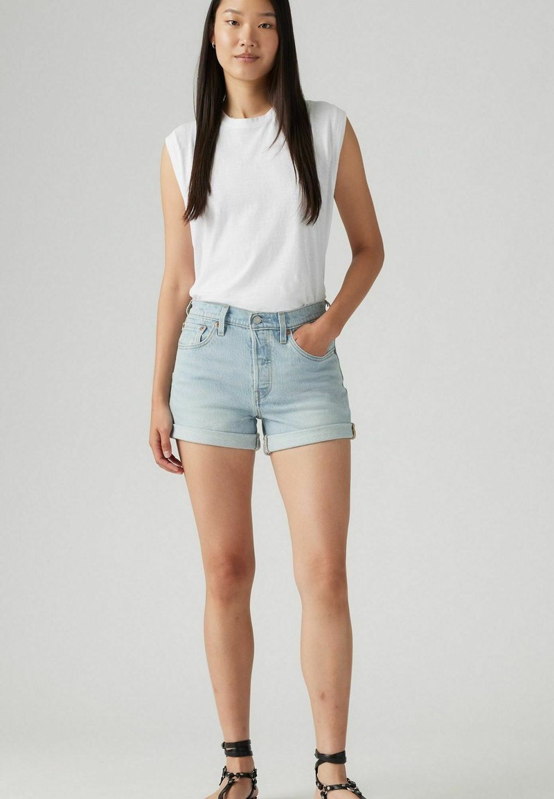 Top blanco sin mangas, pantalones cortos de mezclilla azul claro con puños enrollados, y sandalias negras con tiras. El modelo está de pie con la mano en el bolsillo.