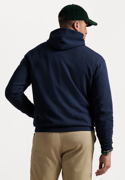 Uomo che indossa un hoodie blu navy, pantaloni beige, un cappello in velluto a coste verde e una camicia a quadri visibile sul polso, di spalle rispetto alla fotocamera.