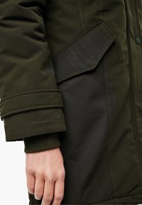 Parka de color verde oliva con una textura suave, que cuenta con un bolsillo con solapa en el brazo, mangas con puños y detalles de costura discretos.
