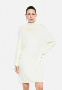 Donna che indossa un vestito a maglia off-white a maniche lunghe con colletto alto e spalle scese, in piedi contro uno sfondo bianco uniforme.