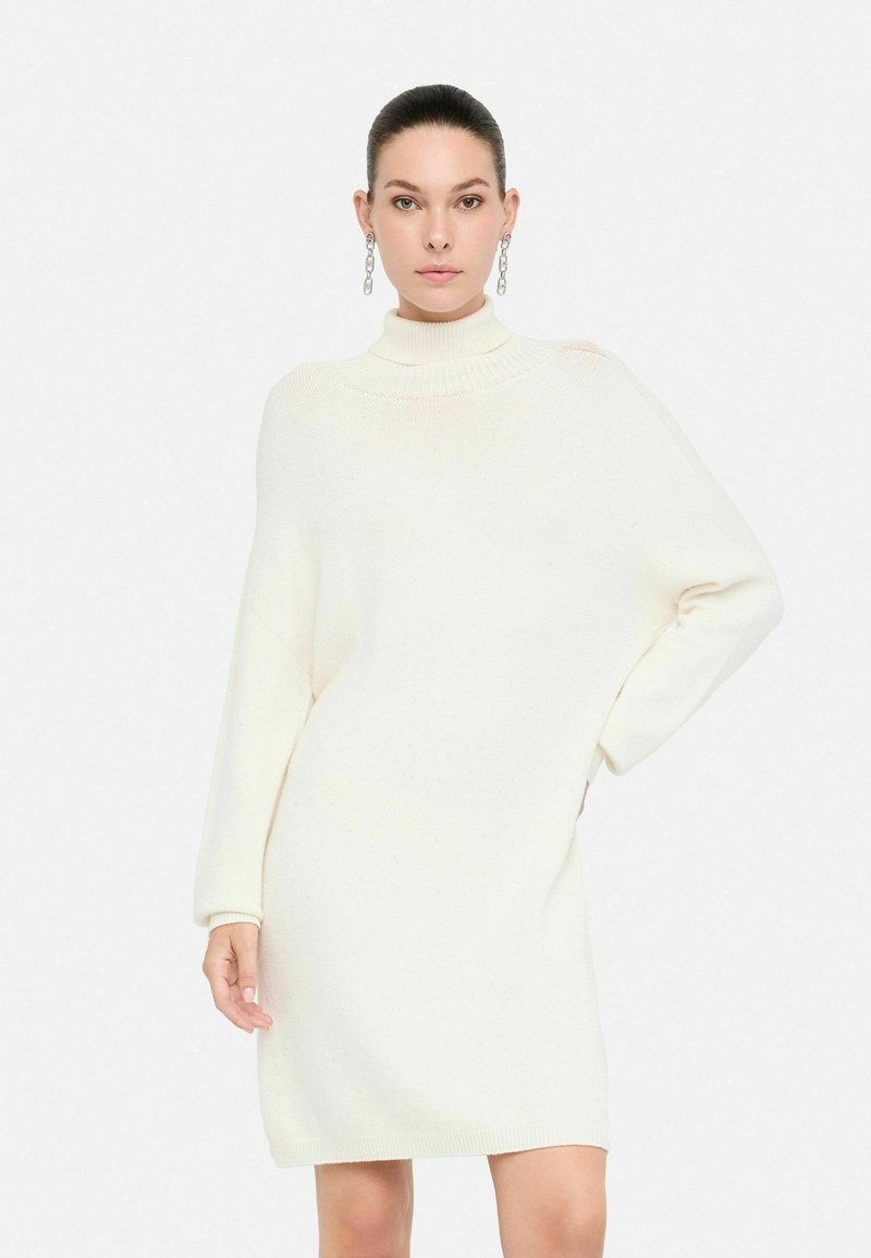 Donna che indossa un vestito a maglia off-white a maniche lunghe con colletto alto e spalle scese, in piedi contro uno sfondo bianco uniforme.