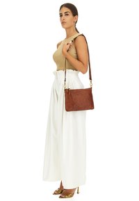Borsa a tracolla in pelle marrone con una texture liscia, hardware dorato e una tracolla marrone, abbinata a un top beige e pantaloni bianchi a gamba larga.