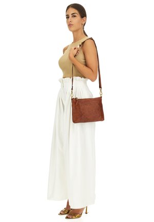 Borsa a tracolla in pelle marrone con una texture liscia, hardware dorato e una tracolla marrone, abbinata a un top beige e pantaloni bianchi a gamba larga.