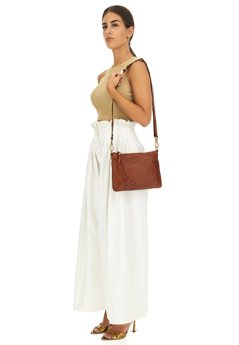Borsa a tracolla in pelle marrone con una texture liscia, hardware dorato e una tracolla marrone, abbinata a un top beige e pantaloni bianchi a gamba larga.