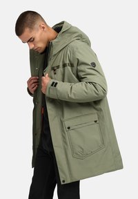 Olivno zelena parka jakna s kapuco, zadrgo in gumbnimi zapiralci, stranskimi žepi ter črnimi poudarki na rokavih. Gladek, vremensko odporen material.