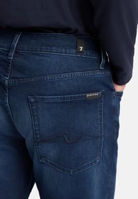 Jeans in denim blu scuro, con vita media, tasche posteriori con cuciture decorative e una piccola etichetta del marchio nera sulla cintura.