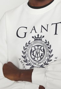 Felpa bianca con scollo rotondo. Presenta un logo grafico navy e un testo, inclusi dettagli con alloro e un disegno di corona. Polsini a coste.