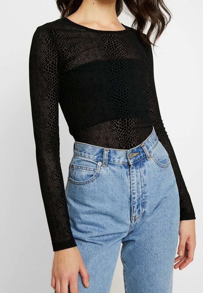 Svart mesh-top med långa ärmar och prickigt mönster, kombinerad med högmidjade ljusblå jeans med knappstängning och fickor.