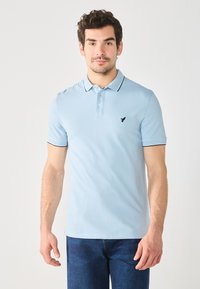 Svijetloplava polo majica s tri gumba, kratkim rukavima i kontrastnim crnim obrubom na ovratniku i rukavima. Mali logo na lijevoj strani prsnog koša.