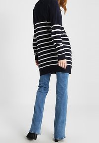 Persona embarazada vestida con un suéter largo de rayas marinas y blancas, con jeans azules y zapatos de tacón negros, de pie frente a un fondo sencillo.