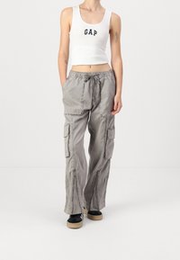 Pantaloni cargo grigi con tasche laterali e dettagli con zip, abbinati a una canotta attillata bianca con logo navy "GAP". Scarpe nere con suole in gomma.