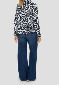 Kvinna klädd i en långärmad skjorta med svart och vit abstrakt mönster och vida blå jeans, stående med ryggen åt kameran.