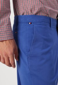 Los pantalones de algodón azul presentan un corte recto y bolsillos laterales sutiles, combinados con una camisa a cuadros roja y negra, que muestra el logo en la cintura.