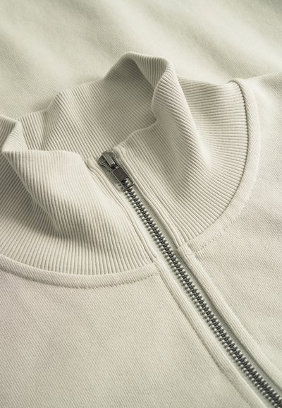 Maglione beige a coste con collo alto e zip in metallo argentato parzialmente chiusa al centro.
