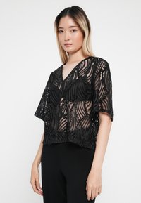 Svart blonde topp med V-hals, korte ermer og intrikate, bølgende mønstre. Stoffet er semitransparent med en teksturert finish.