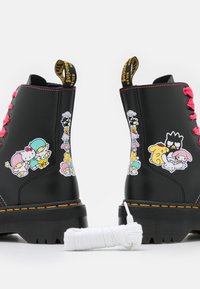 Botas de tobillo de cuero negro con cordones rosas y adhesivos de personajes de dibujos animados coloridos a ambos lados. Suelas gruesas con costuras amarillas.