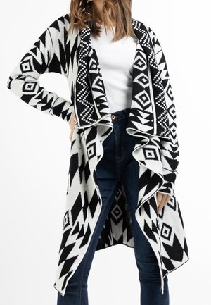 Femme portant un cardigan ouvert à motif géométrique noir et blanc sur une chemise blanche et un jean bleu foncé, debout, la main sur la hanche.