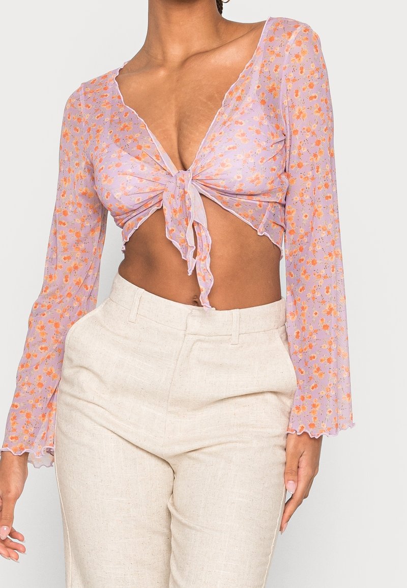 Przezroczysty fioletowy crop top z wzorem kwiatowym, głębokim dekoltem w kształcie litery V i wiązanym przodem; długie rękawy z falbaną. Lekka, zwiewna tekstura.