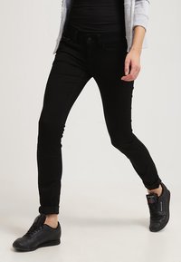 Jeans negros ajustados con una textura suave, con un diseño de cinco bolsillos, cierre de botón y puños enrollados. Combinados con zapatillas negras.