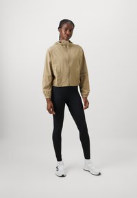 Kvinne står iført en beige jakke med glidelås og hette, svarte leggings, hvite joggesko og hvite sokker mot en enkel lys bakgrunn.