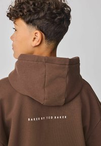 Hnědá mikina s velkou kapucí, vyrobená z texturované látky, na zadní straně s logem "BAKER BY TED BAKER" v bílé barvě.