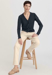 Cardigan gris foncé avec un col en V et des boutons, associé à un pantalon droit beige clair et des mocassins beige, assis sur un tabouret en bois.