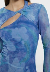 Langärmliges, eng anliegendes Oberteil aus leichtem, transparentem Stoff mit einem blau-grünen Batikmuster. Verfügt über einen einzigartigen gekreuzten Ausschnitt und eine Aussparung.