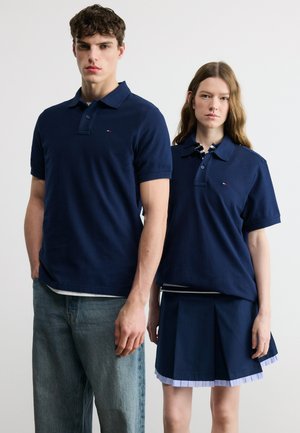 Jonge man en vrouw staan zij aan zij in marineblauwe poloshirts, de man in spijkerbroek, de vrouw in een geplooide rok.