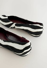 Ballerines à imprimé zèbre noir et blanc avec une texture douce en fourrure, bout arrondi, semelle intérieure rembourrée et semelle extérieure élégante en caoutchouc noir.